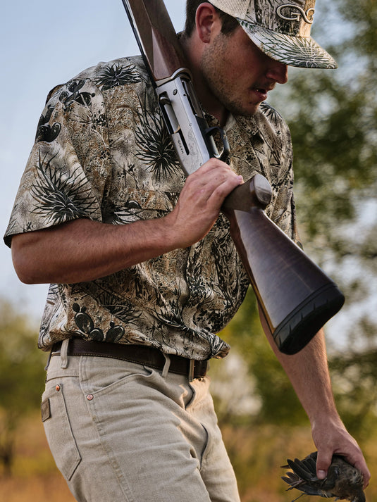 man carrying shotgun in brush and trees wearing gameguard microfiber shirt with dove in hand and gameguard cap with stone meshback